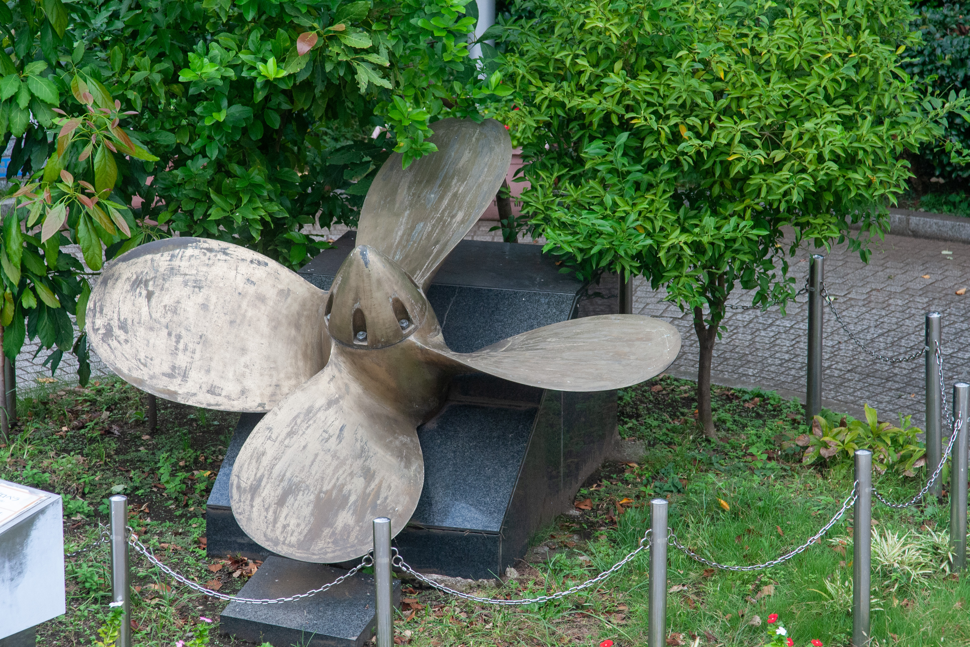 A large used ship propeller is placed on the ground as an ornament surrounded by green trees.