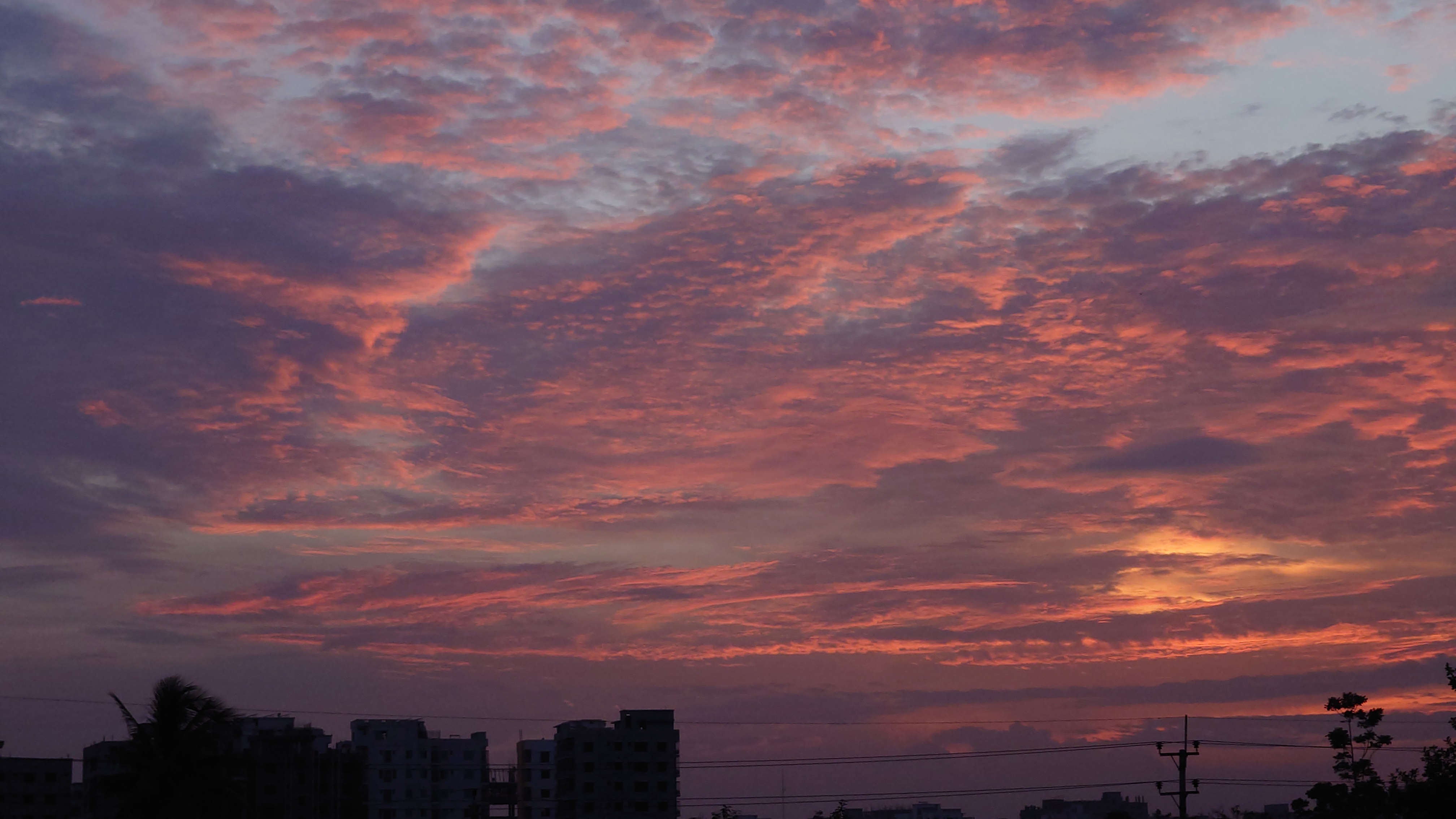 A stunning sunset sky filled with layers of vibrant clouds painted in shades of pink, orange, purple, and blue. The sun is setting below the horizon, casting a warm golden glow that illuminates the lower clouds. Silhouettes of buildings, trees, and power lines appear in the foreground, adding depth and contrast to the colorful evening sky at Dhaka.