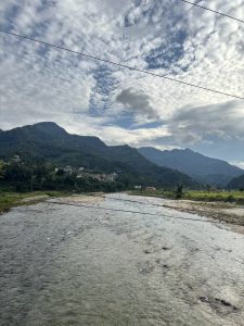 A sceneric view of green hilly background with a flowing river, above is the view of a scattered clouds.