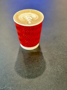A paper cup with latte art on top of hot coffee (cappuccino), placed on a dark tabletop. The warm tones and light shadow add contrast. Shot in Kozhikode, Kerala. 