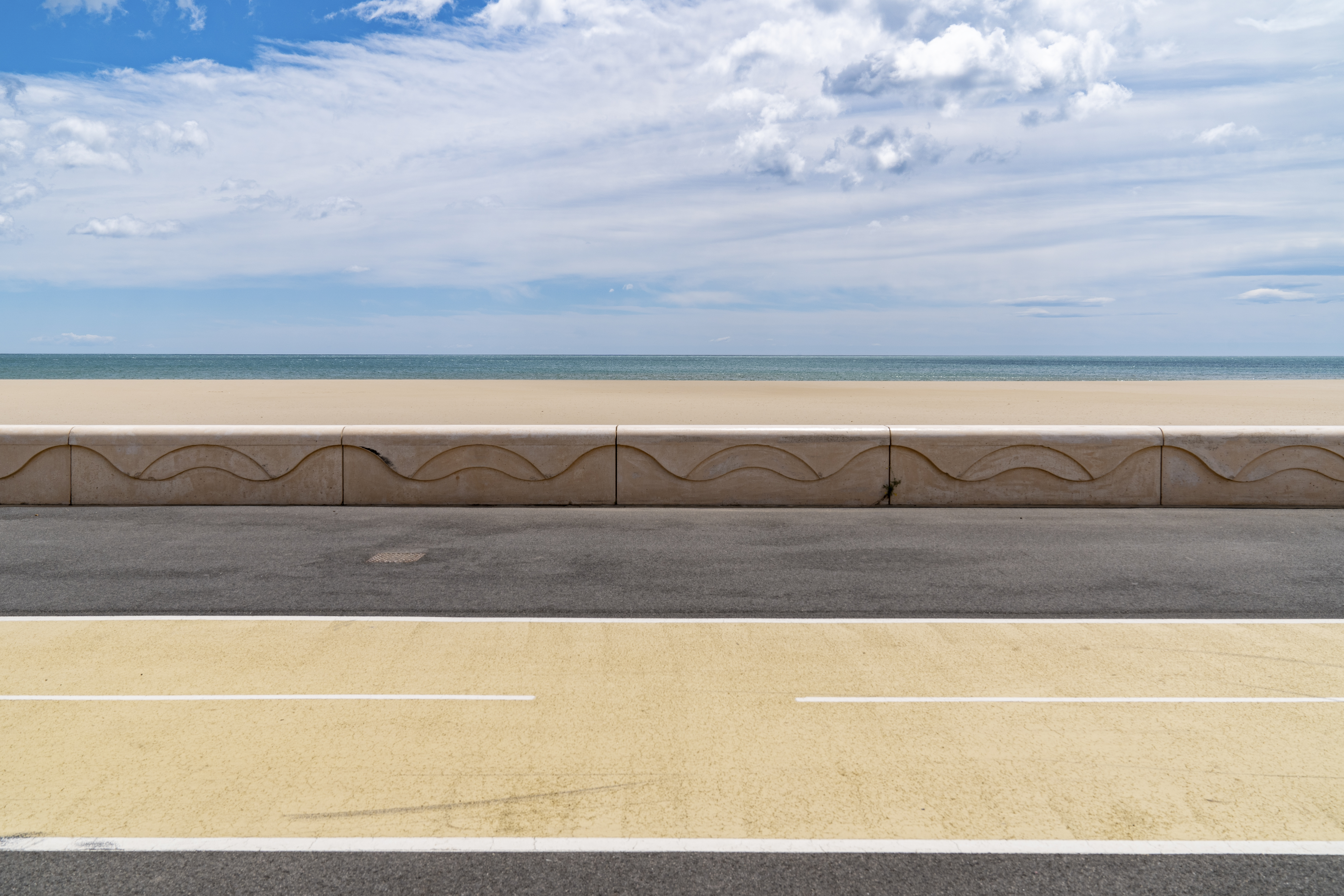 Straight horizon with a minimalistic view of a coastal road, a sandy beach, and the sea under a soft blue sky.