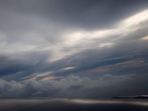 The image features a cloudy sky, filled with various types of clouds creating an atmospheric effect. The landscape appears calm, highlighting the beauty of nature outdoors. The horizon is subtly visible, contributing to the serene scenery that includes elements like water and fog among the cumulus clouds.
