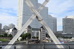 A waterfront cityscape (Yokohama Harbor) framed by intersecting metal beams in the foreground, showing modern high-rise buildings with curved and rectangular shapes. Below, people walk along the water&rsquo;s edge, small boats are docked, and a mix of greenery and urban structures line the shore under a partly cloudy sky.