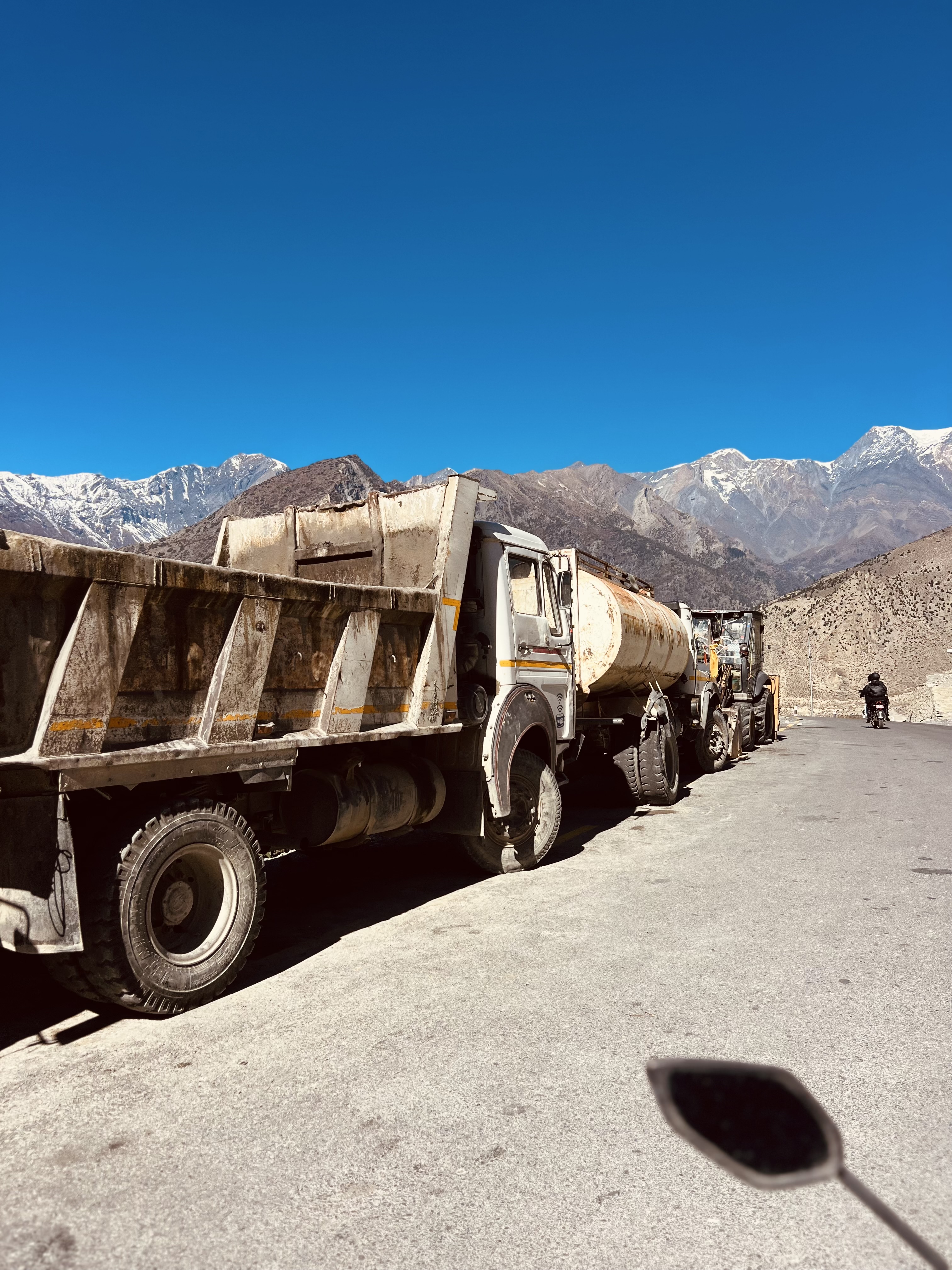 Construction trucks parked on a road with snow-capped mountains in the background.