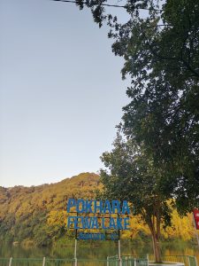 

Fewa Lake in Pokhara, framed by trees and mountains with a blue lakeside sign.