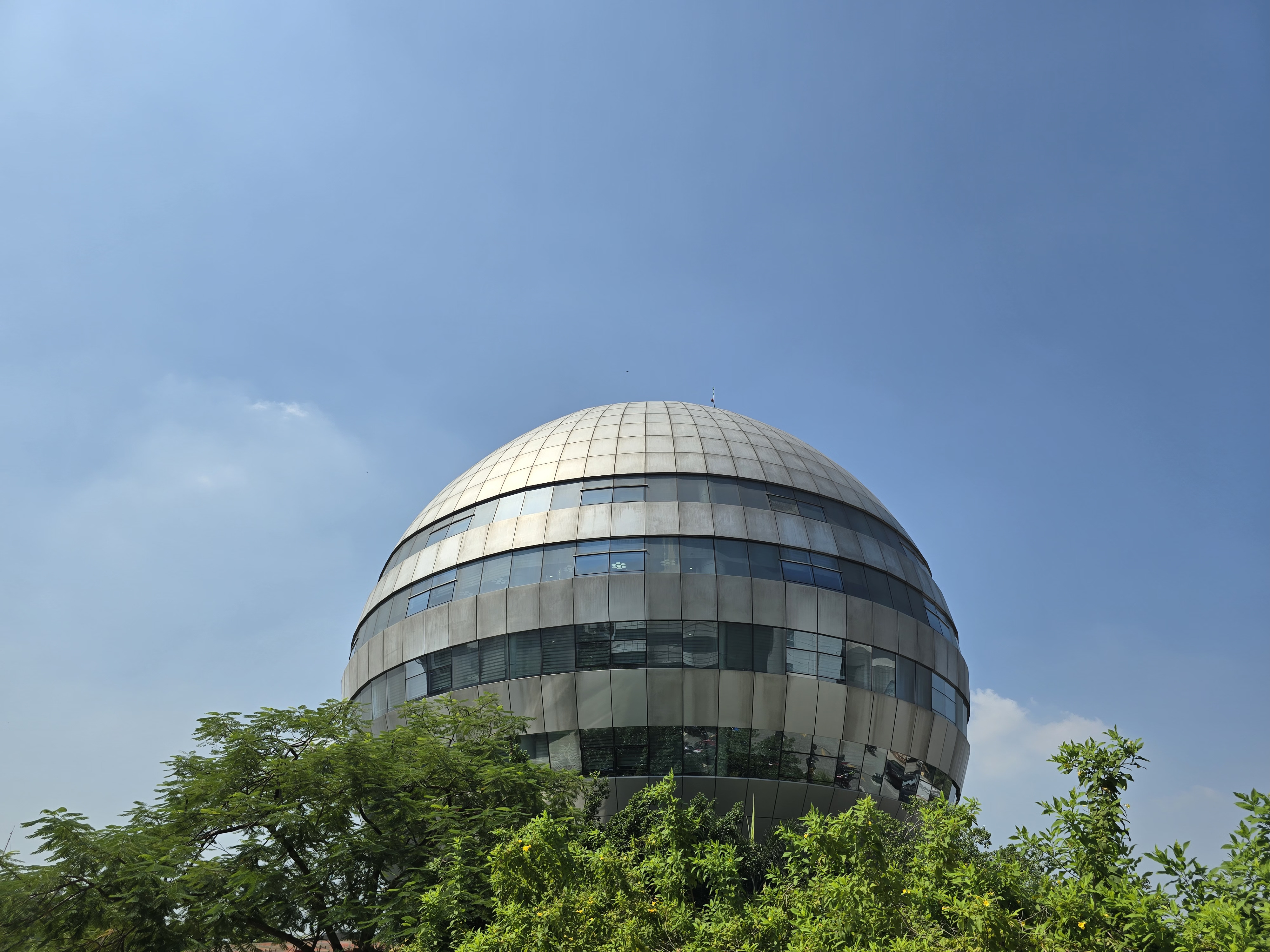 A modern, spherical building with a metallic exterior and large windows, surrounded by green foliage and a clear blue sky.
