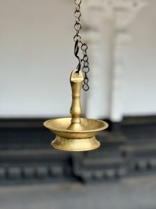 A traditional hanging brass oil lamp (thookku vilakku) suspended by a black metal chain is used for temple rituals. It was photographed at Sree Shiva Vishnu Temple, Perumanna, Kozhikode. 
