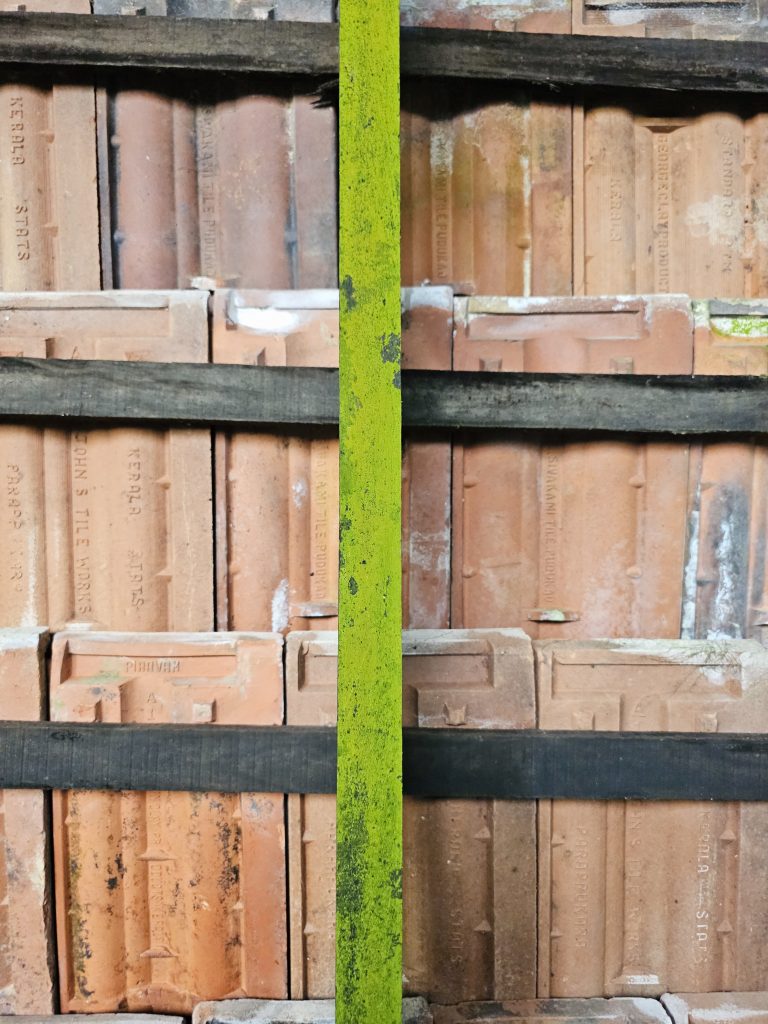 Wooden planks separate a stack of weathered terracotta roof tiles. A bright green, moss-covered pole runs vertically down the center, dividing the image. Taken at Hill Palace, Thrippunithura, Kerala.