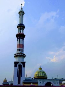 A tall, modern, white, pink, and burgundy minaret of a mosque stands prominently on the left, reaching towards a blue and cloudy sky. To the right is the mosque building, featuring a golden dome with a green and yellow geometric pattern, and an arched entrance with Arabic calligraphy.