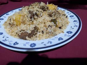 Close-up photograph of a generous serving of Kacchi Biryani, showing spiced long-grain rice, tender chunks of beef, and pieces of potato on a white floral-patterned plate.