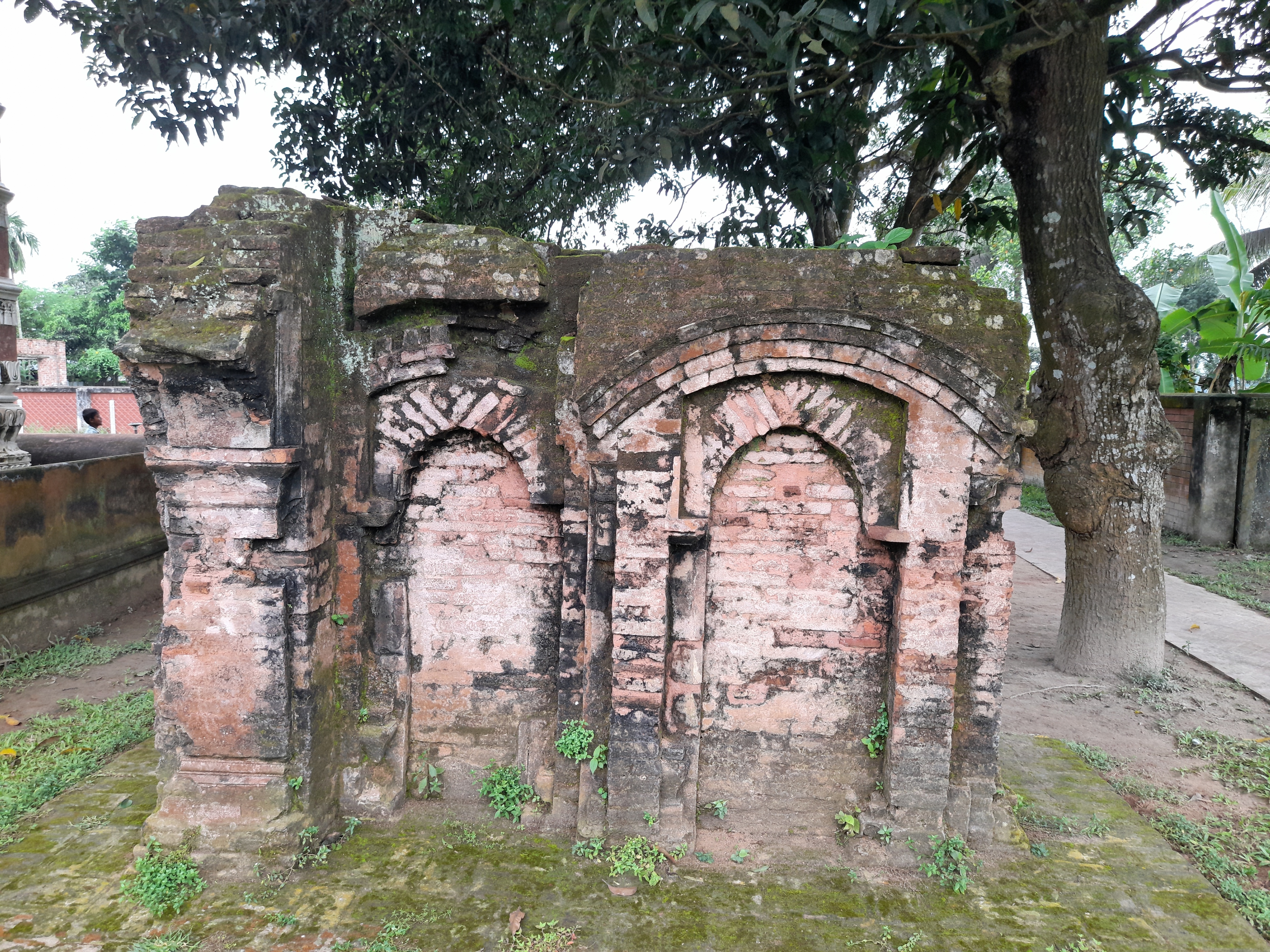 A small moss-covered brick ruin with arched niches stands under a large tree.