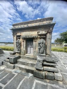 Small shrine at Bucesvara Temple in Koravangala, Hassan, with two guardian sculptures and a wooden door, surrounded by stone steps and paving. 