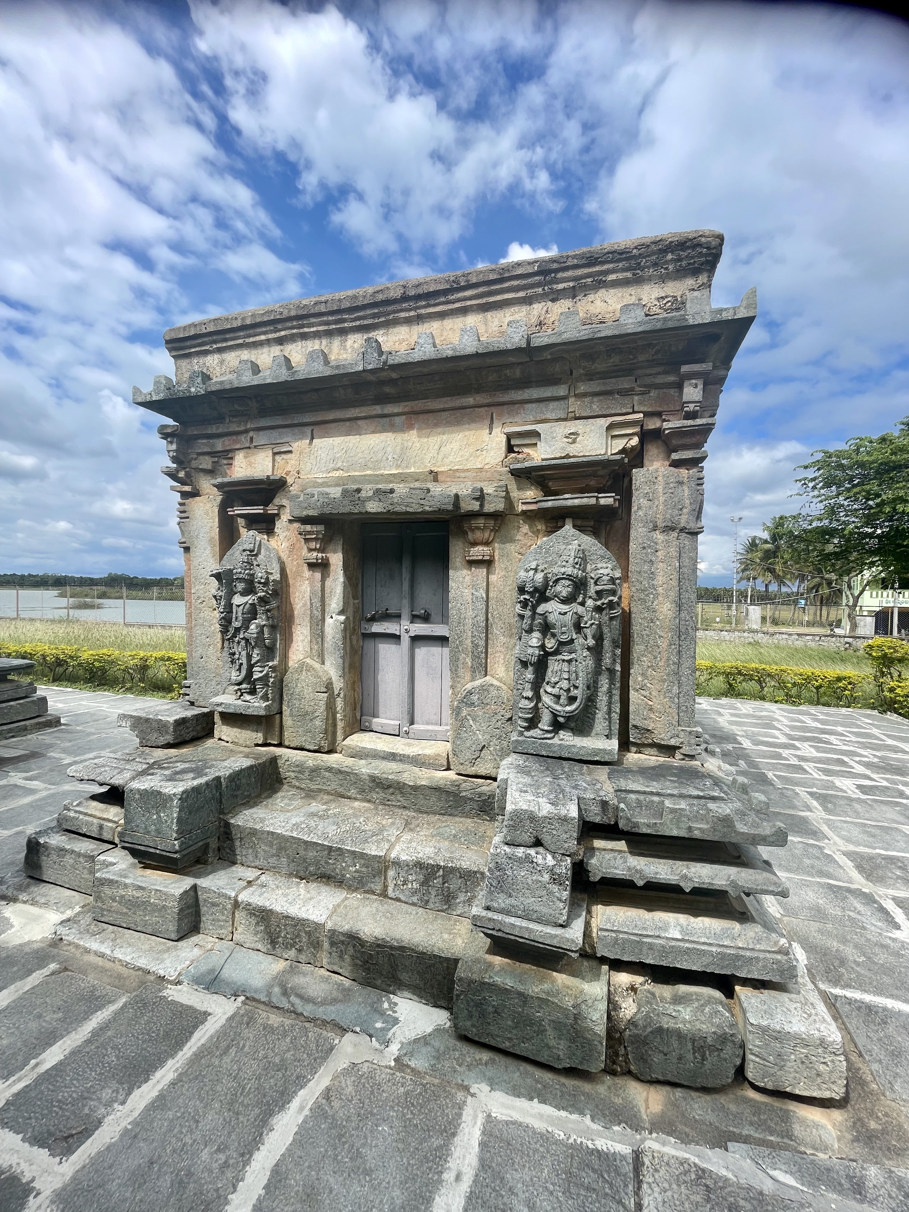 Small shrine at Bucesvara Temple in Koravangala, Hassan, with two guardian sculptures and a wooden door, surrounded by stone steps and paving. 