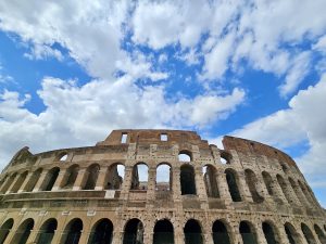 The majestic outer wall of the Colosseum in Rome, Italy, stands tall against a bright blue sky with white clouds, showcasing ancient Roman architecture and historic charm.