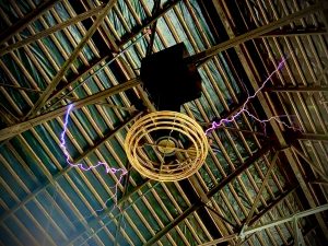 A metal ceiling structure with a large Tesla coil suspended from it, emitting bright purple electrical arcs that extend outward to the surrounding metal beams. The background shows a roof lined with green insulation panels, creating an industrial and electrifying atmosphere.
