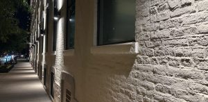 A sidewalk in the left, most of the right side is the wall along the sidewalk with windows, and lights shining down the wall. It&#039;s a night scene.