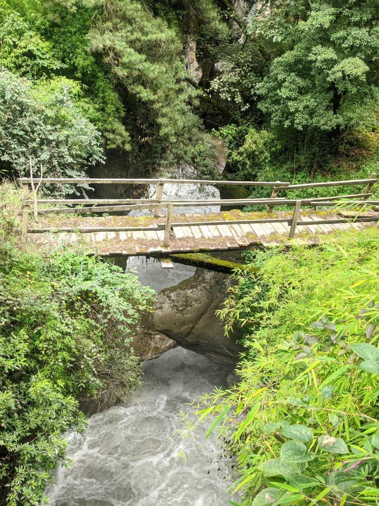 A wooden bridge spans over a flowing river, surrounded by lush greenery and rocky formations.