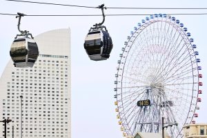 Two cable cars pass by a colorful Ferris wheel and a curved building in Yokohama, Japan, under a clear sky.
