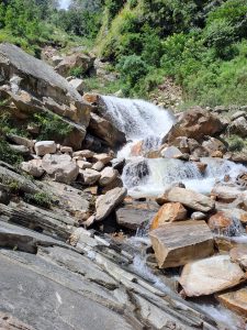 A cascading waterfall flows over large, rugged rocks surrounded by lush greenery. 