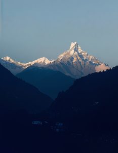 A majestic snow-capped Fishtail mountain peak rises prominently against a clear sky, flanked by rolling hills and a mountain range in the foreground.
