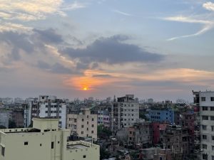 A city skyline at sunset with warm orange and yellow hues in the sky, scattered clouds, and a mix of older colorful buildings in the foreground and modern high-rises in the background.