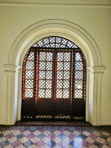 An arched wooden window with an ornamental grill lets sunlight into a hallway at Hill Palace, Thrippunithura, Kerala. The old architecture adds timeless charm. 