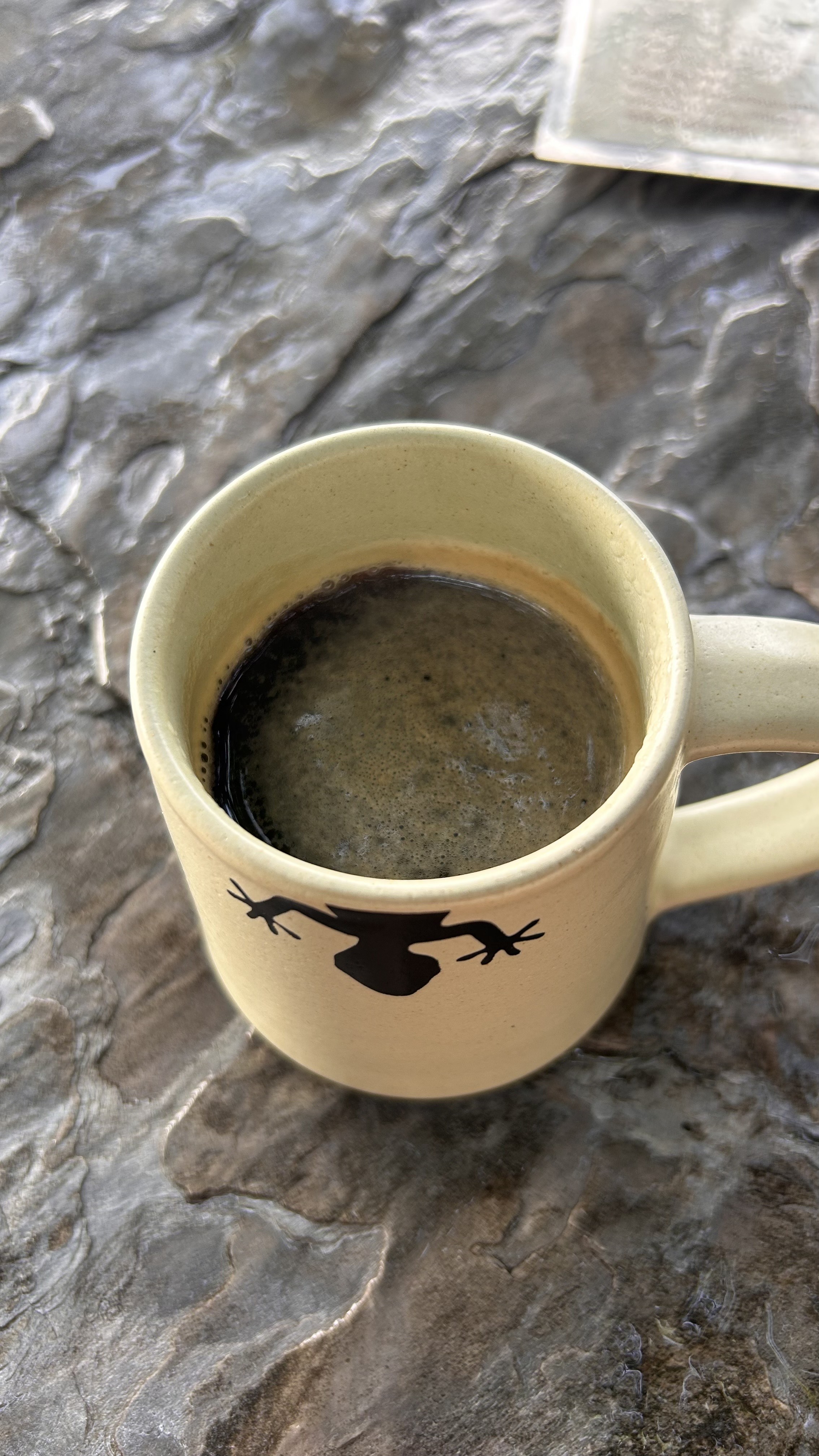 A close-up view of a beige mug with a frog silhouette on the side, filled with dark coffee.