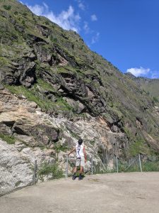 A person stands at the edge of a rocky cliff, looking up at a steep mountain covered in greenery under a bright blue sky with a few clouds.