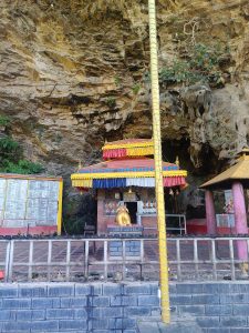 

A small Changchang di temple sanyanja structure is located at the base of a rocky cliff, surrounded by greenery. 