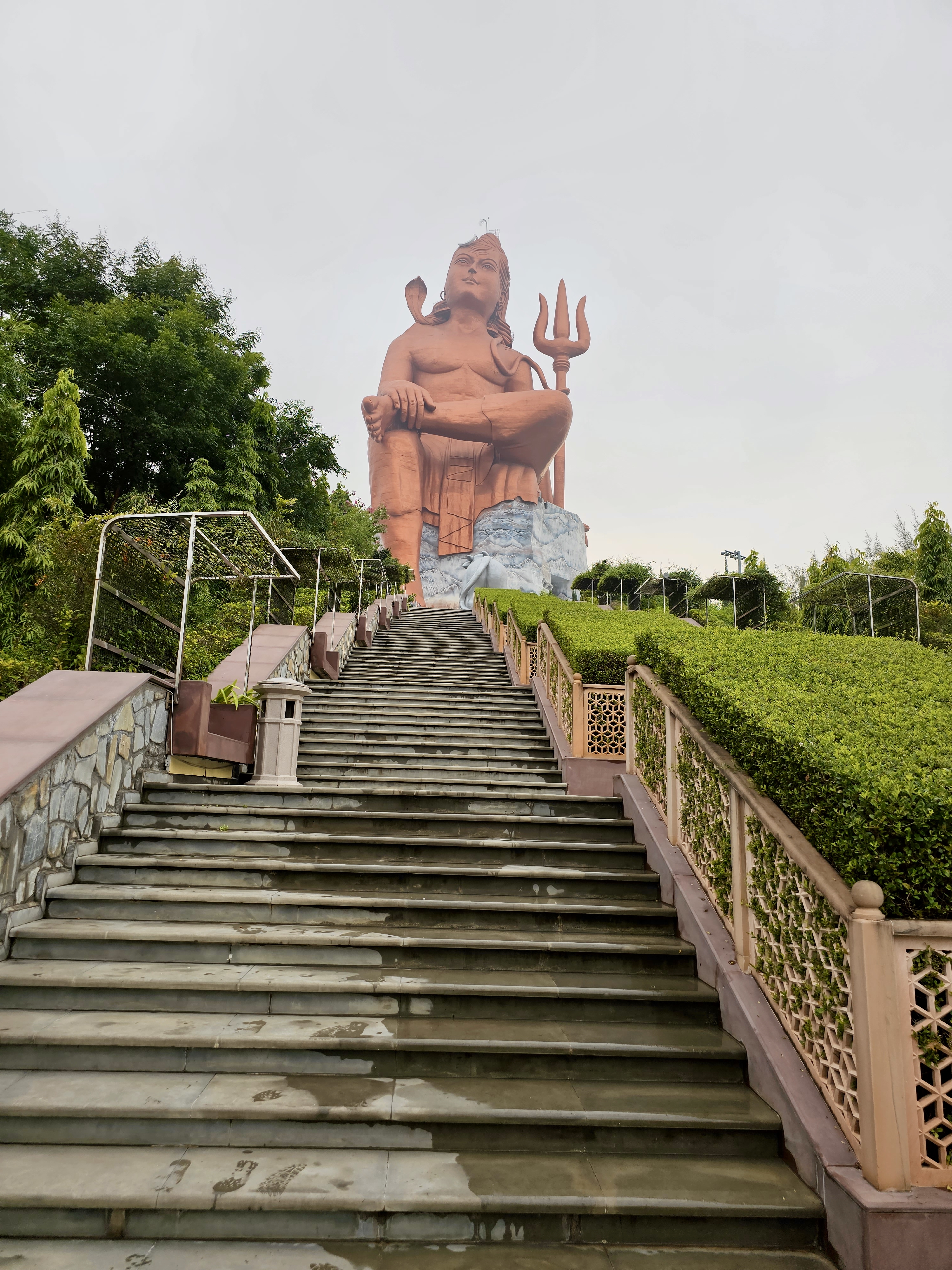 A large Shiva statue (Statue of Belief, aka Vishwas Swaroopam) at the top of a staircase in Nathdwara, Rajasthan. The clean stone steps lead to the tall idol of Lord Shiva with a trident, surrounded by greenery. 