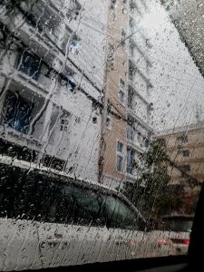 Raindrops on a car window blur the view of tall buildings and a parked white car outside. The scene conveys a moody, rainy day atmosphere at Uttara, Dhaka.

