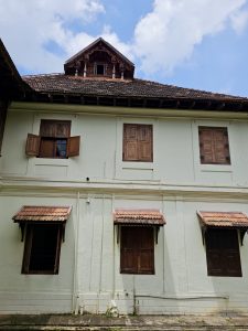 A traditional Kerala palace building with wooden windows and tiled roof at Hill Palace, Thrippunithura, Kerala. The photo shows a mix of colonial and local architecture set against a blue sky. 