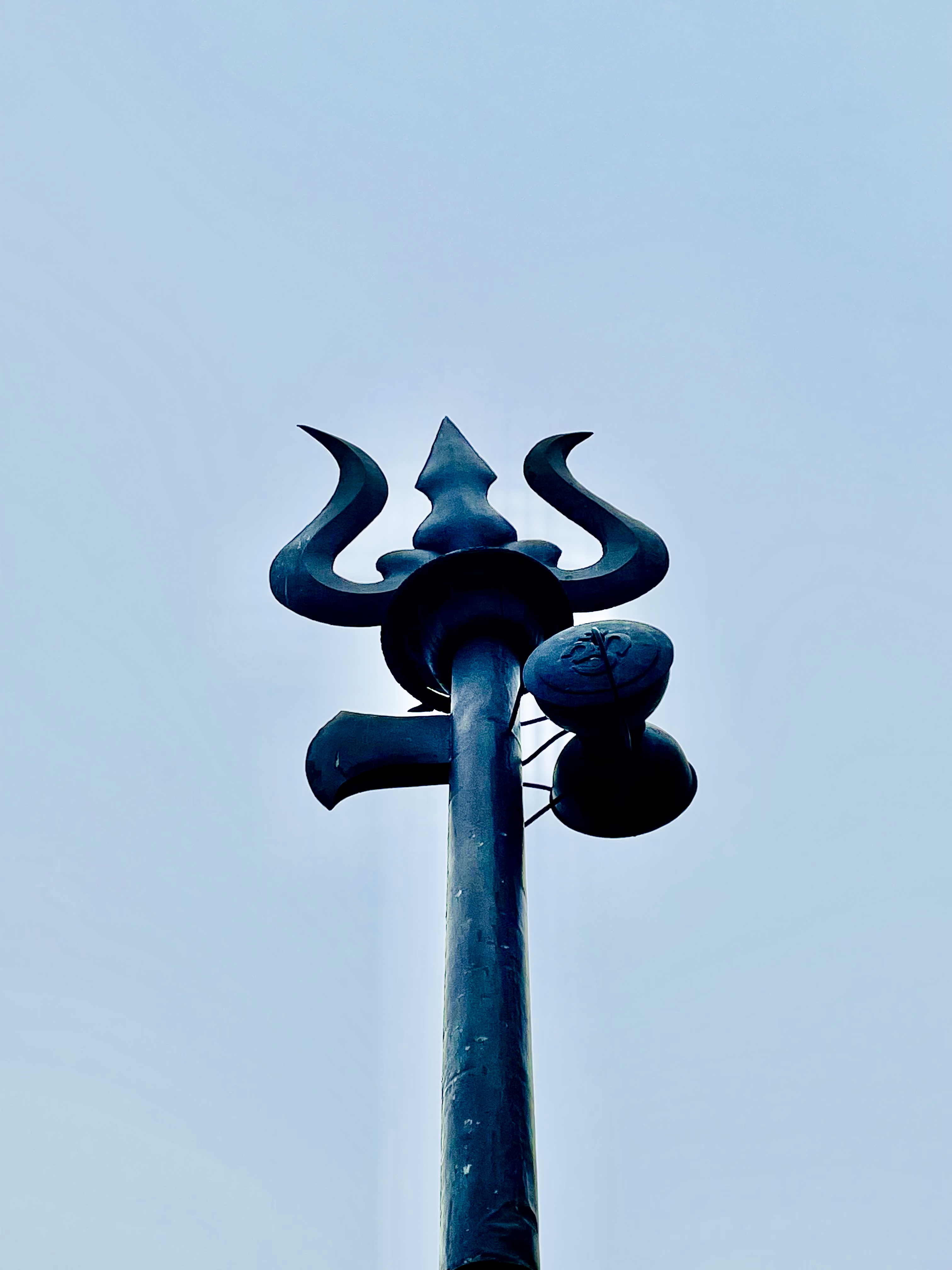 A dark Trishul (trident) with a damaru attached to a tall pole is photographed from below against a clear sky at Bhaleshwor Mahadev Temple, Chandragiri Hill, Nepal. 