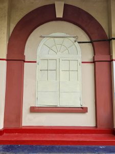 An arched, vintage-style window painted in pale yellow with a red border, located on an old wall. This is part of the abandoned Cochin Harbour Terminus in Kochi. 