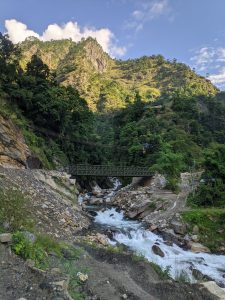 A scenic view depicting a mountainous landscape with lush greenery and rocky terrain. In the foreground, a river flows swiftly over stones, while a small, green metal bridge spans the river.
