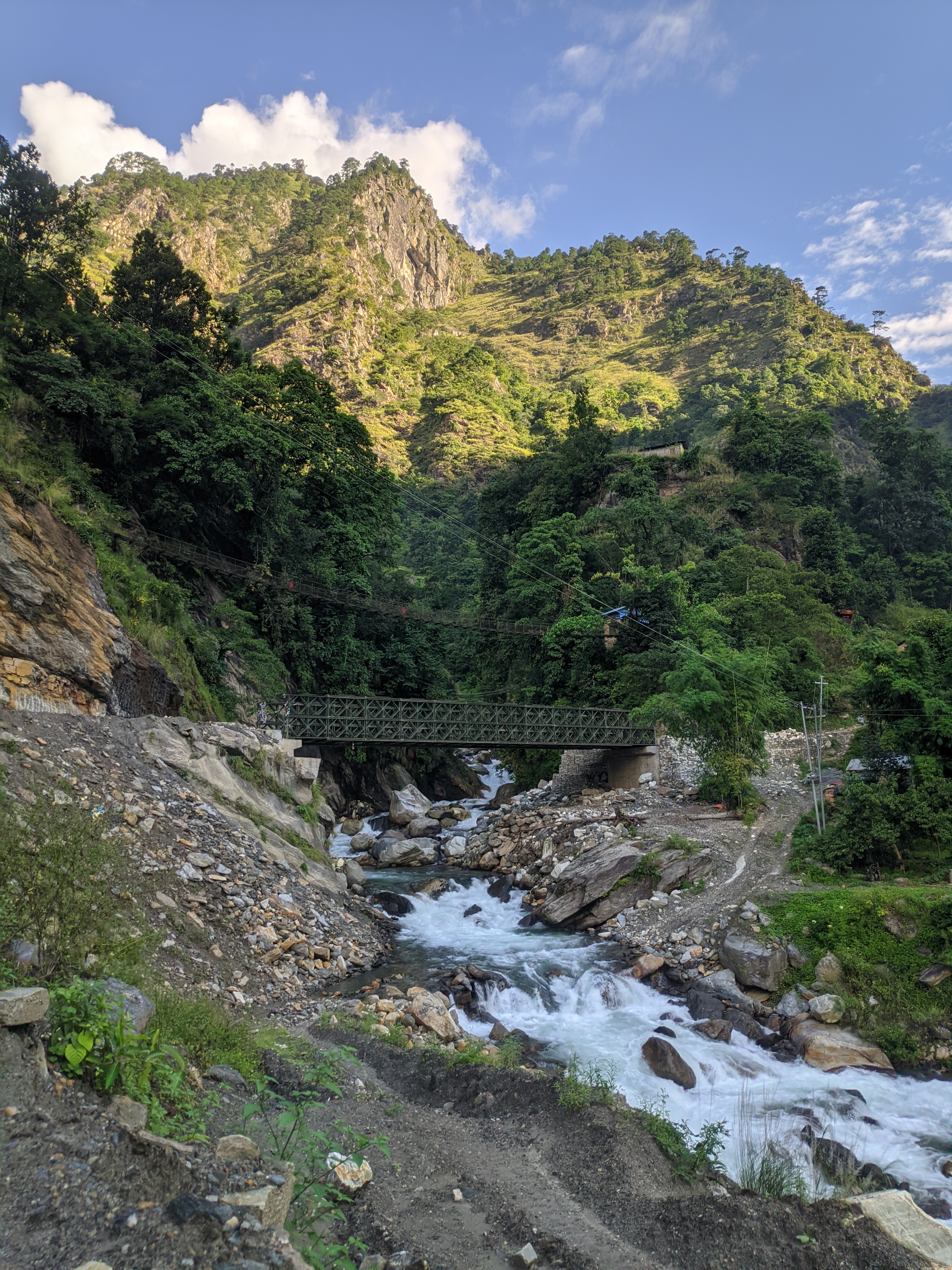 A scenic view depicting a mountainous landscape with lush greenery and rocky terrain. In the foreground, a river flows swiftly over stones, while a small, green metal bridge spans the river.
