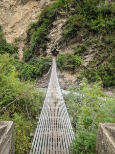 A narrow suspension bridge made of metal grating stretches across a rocky landscape, surrounded by lush greenery. 