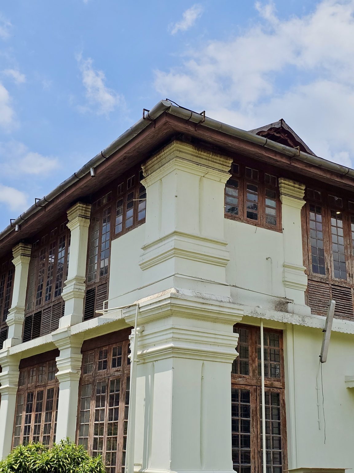 A close-up view of the top floor of the Hill Palace museum in Thrippunithura, showing tall wooden windows, thick white pillars, and a tiled roof under a blue sky.