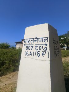 A white concrete border pillar marking the boundary between India and Nepal under a clear blue sky. The text on the pillar is written in Devanagari script, with arrows pointing in opposite directions — one side labeled "भारत नेपाल" (India–Nepal) with numbers “807 (6A)” and the other labeled "नेपाल" (Nepal). The pillar stands in a grassy rural area with a small building and trees in the background.