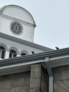 Close-up of a building roof with a white curved structure and round window, two green parrots perched on the edge, gray stone wall below, under an overcast sky.