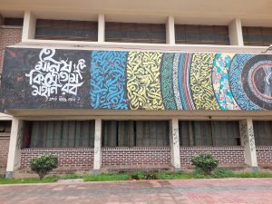 A colorful mural with intricate patterns and Bengali calligraphy adorns a brick building, flanked by two small bushes.