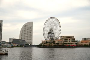 A large Ferris wheel stands beside a uniquely shaped modern building on an overcast day, its reflection shimmering on the calm waterfront with a pier in the foreground.