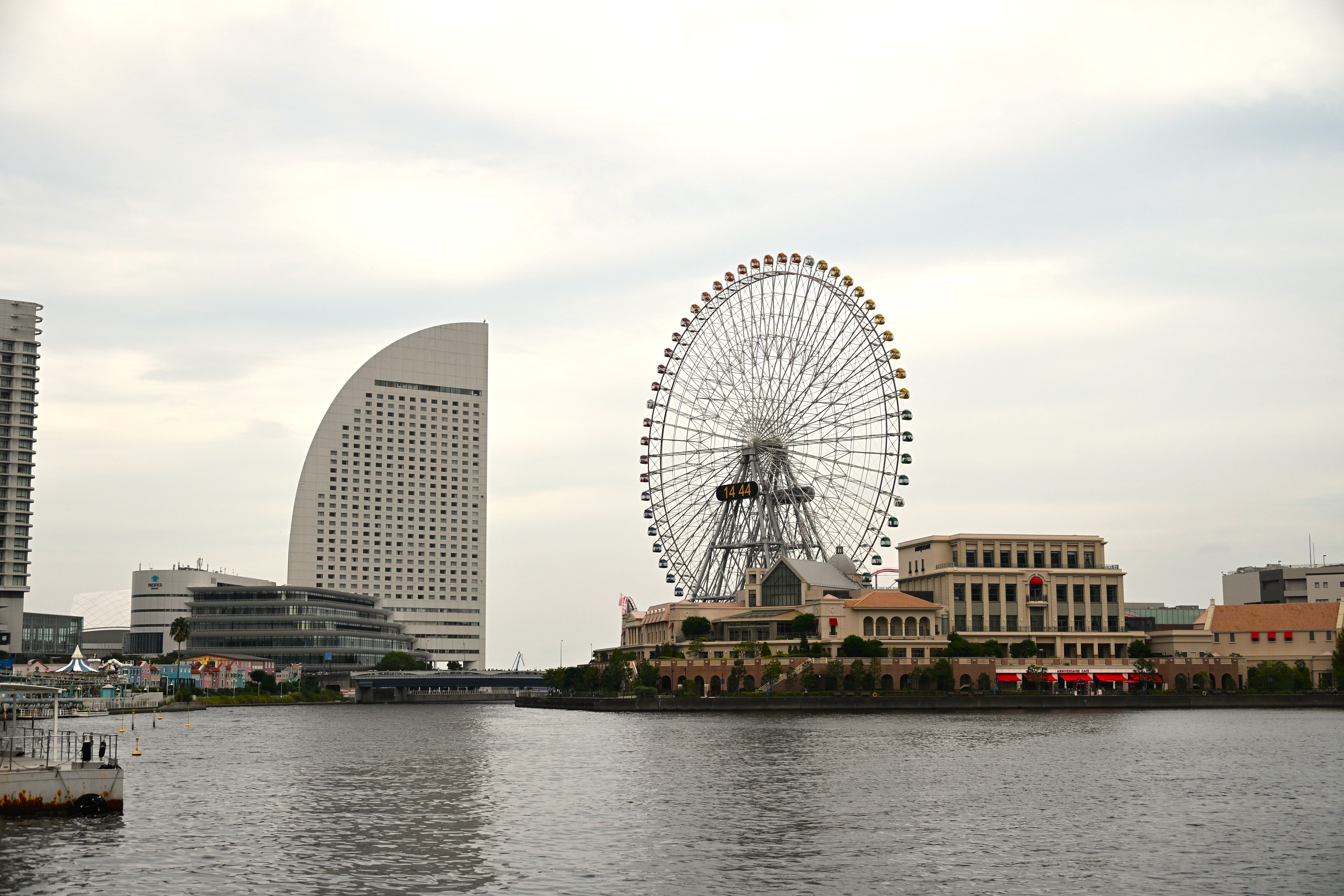 A large Ferris wheel stands beside a uniquely shaped modern building on an overcast day, its reflection shimmering on the calm waterfront with a pier in the foreground.
