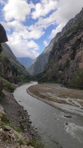 The image shows a stunning mountain range featuring a river flowing through the bottom. The upper view had beautiful cloudy sky. The river banks are rocky with some dried grasses and sand.