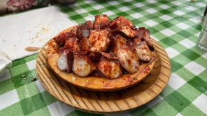 Plate of &ldquo;pulpo &aacute; feira&rdquo;: sliced octopus drizzled with olive oil and sprinkled with paprika on a round wooden dish, set on a green-and-white checkered tablecloth with a toothpick in the center.
