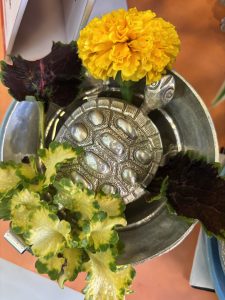 A silver decorative turtle-shaped container sits in the center of an arrangement, surrounded by vibrant green and purple leafy greens, and a bright yellow marigold flower.