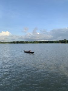 A lone fisherman in a small wooden boat floating on a calm river under a clear blue sky with scattered clouds and green trees in the background