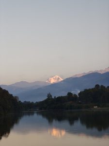 A calm Fewa lake reflects the surrounding landscape, with mountains in the background partially covered by light clouds.