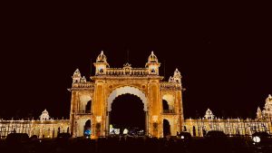Mysore palace gate. A grand illuminated entrance archway adorned with thousands of lights, set against a dark night sky. The architecture features intricate designs with domes and arches, showcasing a festive atmosphere. In the foreground, a crowd of people can be seen gathering near the entrance.
