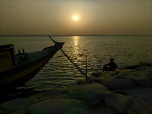 A beautiful scene of the river with sunset view. A man fishing on the edge of the river is visible along with visibility of some rocks and a boat.